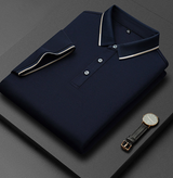 Camicia polo blu scuro piegata con colletto a contrasto, abbinata a un elegante orologio da polso nero su sfondo grigio. Moda uomo, abbigliamento casual.