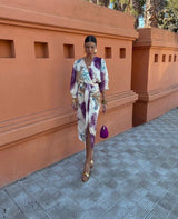 Donna con abito floreale elegante, sandali dorati e borsa viola, cammina su marciapiede davanti a muro arancione. Moda estiva, stile sofisticato.