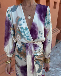 Abito elegante donna con stampa astratta multicolore, scollo a V profondo, maniche lunghe, cintura in vita, accessori oro, sfondo arancione.