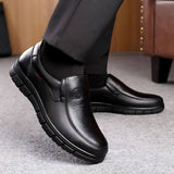 Scarpe eleganti da uomo in pelle nera, design slip-on, suola antiscivolo, indossate con pantaloni scuri su pavimento grigio.