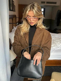 Donna bionda con occhiali rosa indossa giacca marrone e maglione nero, tiene borsa nera in camera da letto moderna. Moda autunnale elegante.